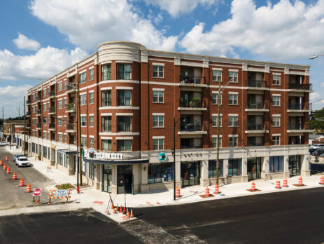 Aerial view of a multi-story condo building in Berwyn, IL, captured by a drone. The image highlights the building’s brick façade and balconies, with a clear view of its rooftop and surrounding area. Below, you can see neatly arranged parking spaces, landscaped pathways, and adjacent residential homes. The photo captures the urban-suburban mix of Berwyn, with tree-lined streets and nearby small businesses visible in the background, all under a sunny, cloud-dotted sky.