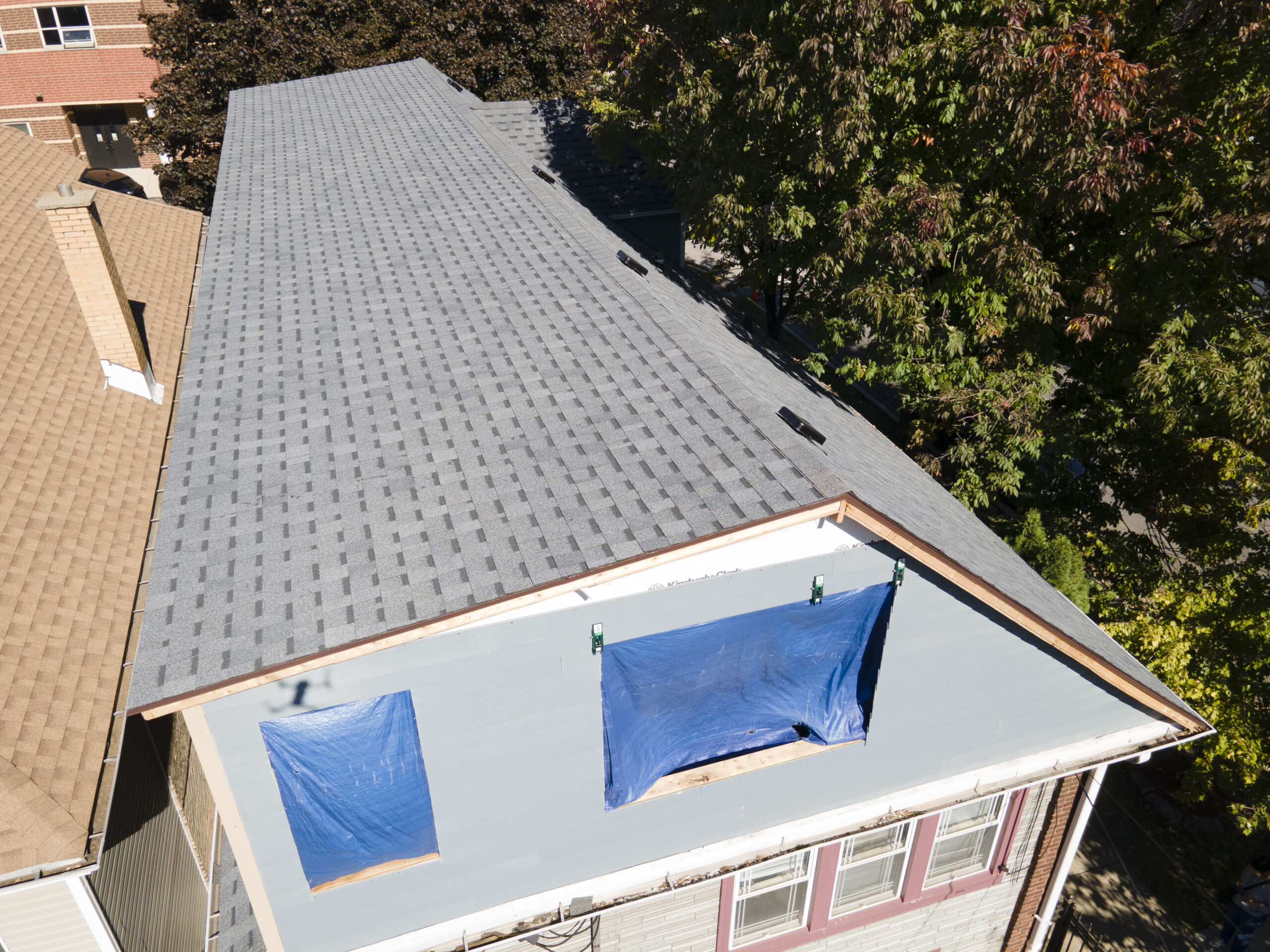 Aerial view of a roof construction job in progress. The image captures workers in safety gear positioned along the roof structure, laying new shingles and installing roofing materials. Scaffolding and ladders surround the building, and various construction tools and supplies are spread out on the ground below. The roof is partially completed, revealing both the exposed underlayment and newly installed sections. Nearby, parked trucks and equipment signal an active job site. The setting is a suburban neighborhood with well-kept lawns and houses in the background, framed by a sunny sky overhead.