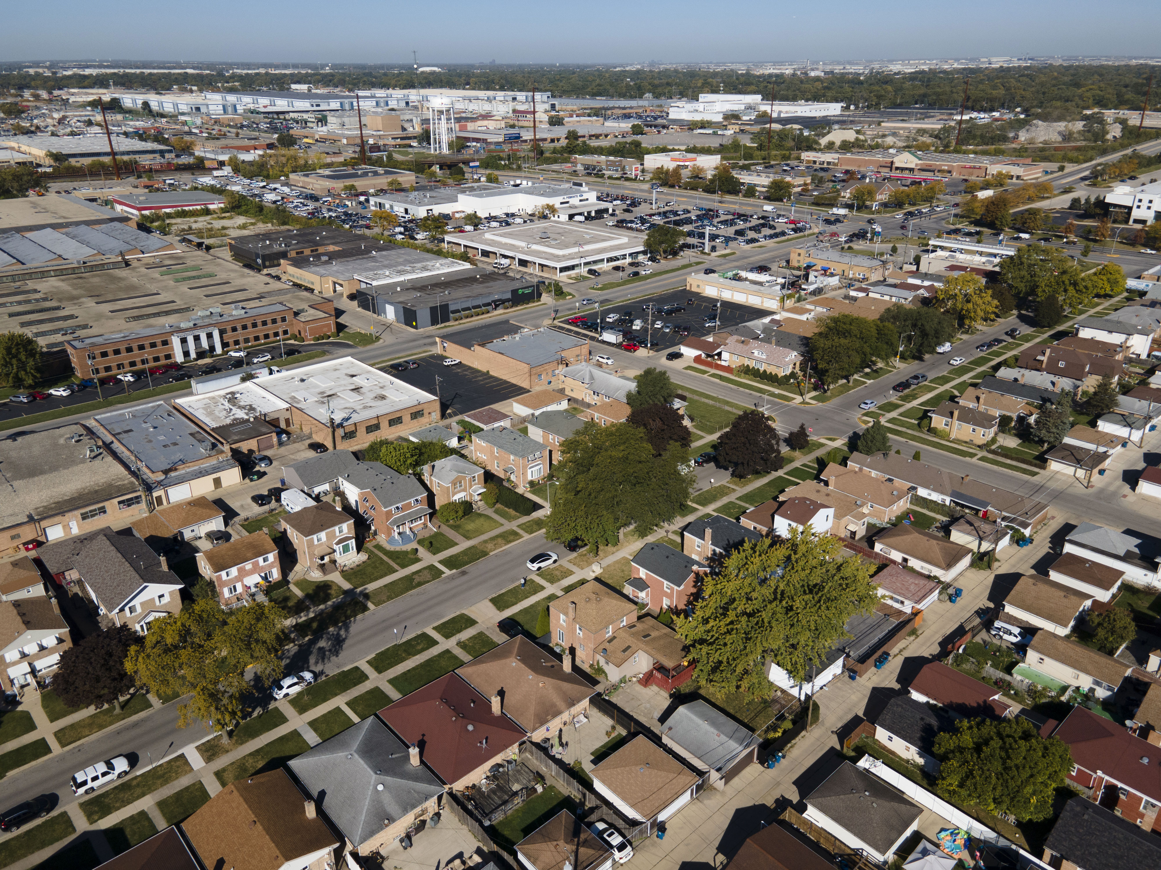 Aerial view of Melrose Park, IL, captured by a drone. The image shows a mix of residential, commercial, and industrial areas, with tree-lined streets dividing rows of brick houses and small businesses. The industrial zone features large warehouses and factory buildings with visible rooftops and parking lots. In the center, a prominent park space with sports fields and greenery breaks up the urban landscape. The scene is framed by major roadways, and cars are seen moving along, highlighting the area's connectivity. The sky above is clear, giving the town a bright, spacious look.